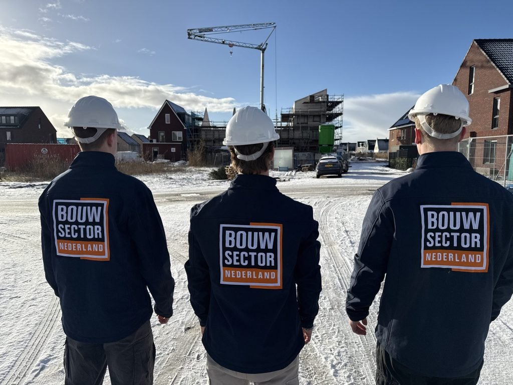 Drie vakmensen van Bouwsector Nederland met witte helm op een besneeuwde bouwplaats met rijwoningen in aanbouw en een bouwkraan